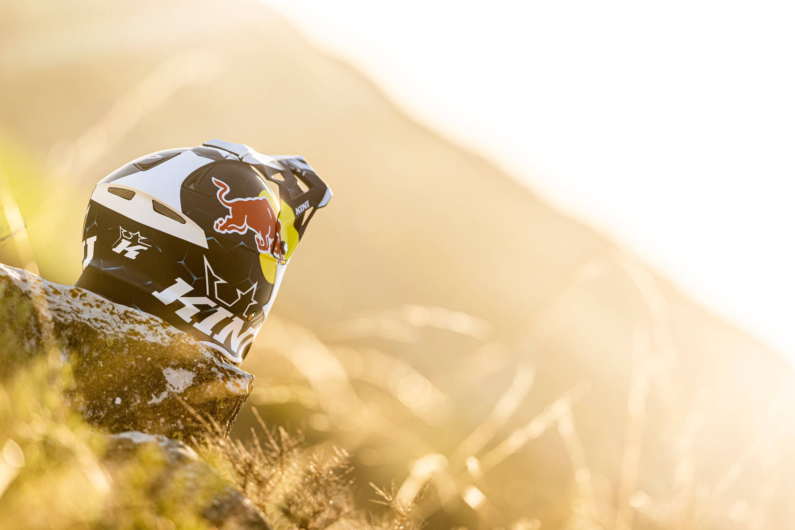 Ein Motocross-Helm mit Red Bull- und 100%-Logos ruht auf einem Felsen im goldenen Sonnenlicht, umgeben von hohem trockenem Gras und einem sanft verschwommenen natürlichen Hintergrund.